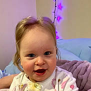 Serenity is registered to the contest to win money with this photo: child, baby, toddler, face, smile, blue_eyes, pajamas, onesie, blanket, pillow, bed, indoor, close_up, portrait, hair, rosy_cheeks, string_lights, purple_lights, wall, happy