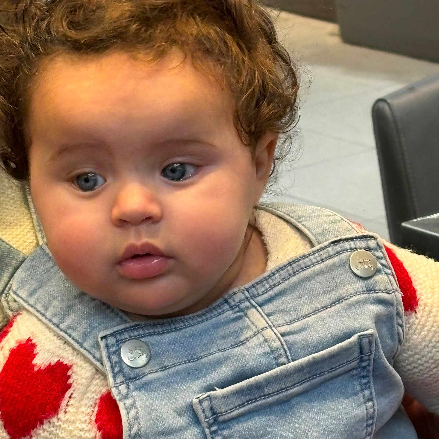 Lyna participe au concours pour gagner de l'argent avec cette photo : baby, child, face, curly_hair, blue_eyes, denim_overalls, sweater, red_hearts, indoor, seated, cute, portrait, soft_lighting, chubby_cheeks, person, clothing, expression, closeup, infant, casual