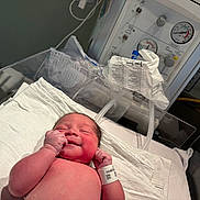Avery is registered to the contest to win money with this photo: newborn, baby, hospital, medical_equipment, bassinet, infant, skin, wrinkled_skin, wristband, healthcare, bed, white_towel, smiling, cute, fresh, person, patient, new_life, indoor, closeup