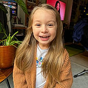 Olivia a rejoint le concours — aidez-le/la à gagner de superbes lots ! child, smiling, long_hair, cardigan, indoor, plant, happy, portrait, person, living_room, furniture, cozy, casual_clothing, young, blonde, cute, home, sitting, natural_light, warm_colors