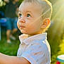 toddler, child, portrait, outdoor, sunlight, golden_hour, curly_hair, white_shirt, patterned_clothing, greenery, sunflare, face, looking_up, cute, young_child, short_sleeves, daylight, background_blur, nature, innocence