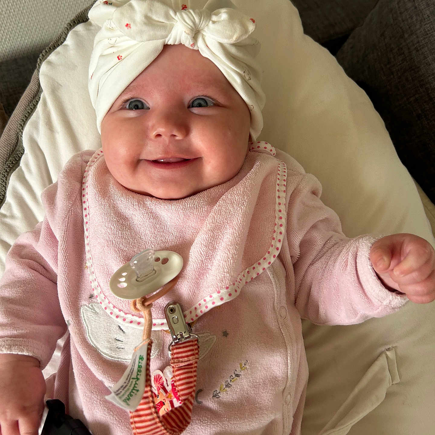 Giulia a rejoint le concours — aidez-le/la à gagner de superbes lots ! adorable, baby, blanket, child, closeup, comfort, cushion, cute, face, hands, happy, head_wrap, indoors, infant, pacifier, pink_clothing, portrait, seated, smiling, soft_toys
