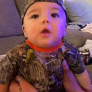 Ryland is registered to the contest to win money with this photo: baby, child, camo_clothing, cap, backward_cap, tattooed_hands, sofa, couch, living_room, purple_light, curious_expression, person, indoor, casual_clothing, seated, holding, cute, wide_eyes, infant, young_child