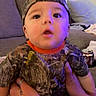 baby, child, camo_clothing, cap, backward_cap, tattooed_hands, sofa, couch, living_room, purple_light, curious_expression, person, indoor, casual_clothing, seated, holding, cute, wide_eyes, infant, young_child