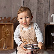 Sacha participe au concours pour gagner de l'argent avec cette photo : child, baby, toddler, one_year, first_birthday, smiling, portrait, indoor, wooden_background, crate, steering_wheel, knit_overall, long_sleeve, sitting, props, cotton_branch, vase, number_one, neutral_colors, studio_photo