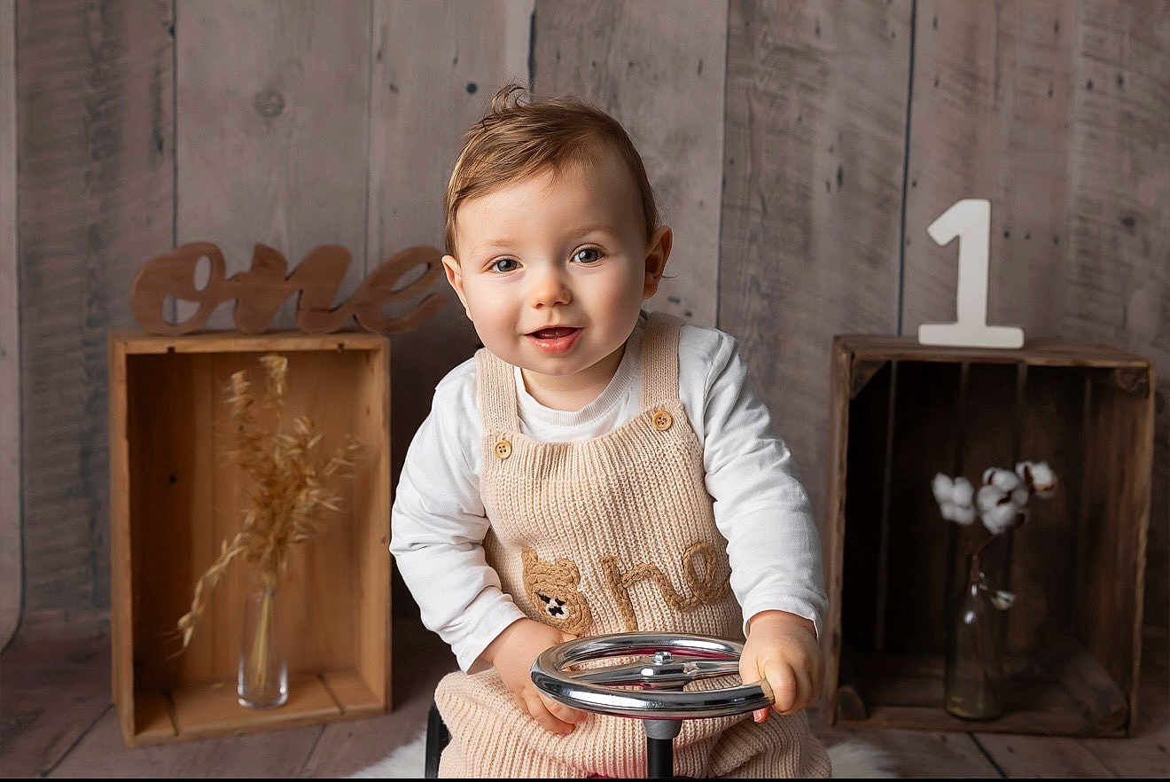 Sacha participe au concours pour gagner de l'argent avec cette photo : child, baby, toddler, one_year, first_birthday, smiling, portrait, indoor, wooden_background, crate, steering_wheel, knit_overall, long_sleeve, sitting, props, cotton_branch, vase, number_one, neutral_colors, studio_photo