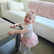 Layla is registered to the contest to win money with this photo: baby, bow, carpet, child, couch, cute, hair_accessory, happy, home, indoor, living_room, person, pillow, pink_tutu, smiling, standing, table, toddler, white_shirt, young_child
