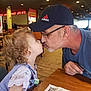 child, toddler, man, kiss, restaurant, table, curly_hair, cap, glasses, wooden_floor, menu, indoor, casual_clothing, playful, love, family, interaction, people, seated, light