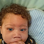 Nasir is registered to the contest to win money with this photo: toddler, child, face, hands, curly_hair, blue_eyes, cushion, checkered_pattern, indoor, portrait, casual_clothing, baby, cute, young_child, hand_in_mouth, seated, expression, person, close_up, soft_background