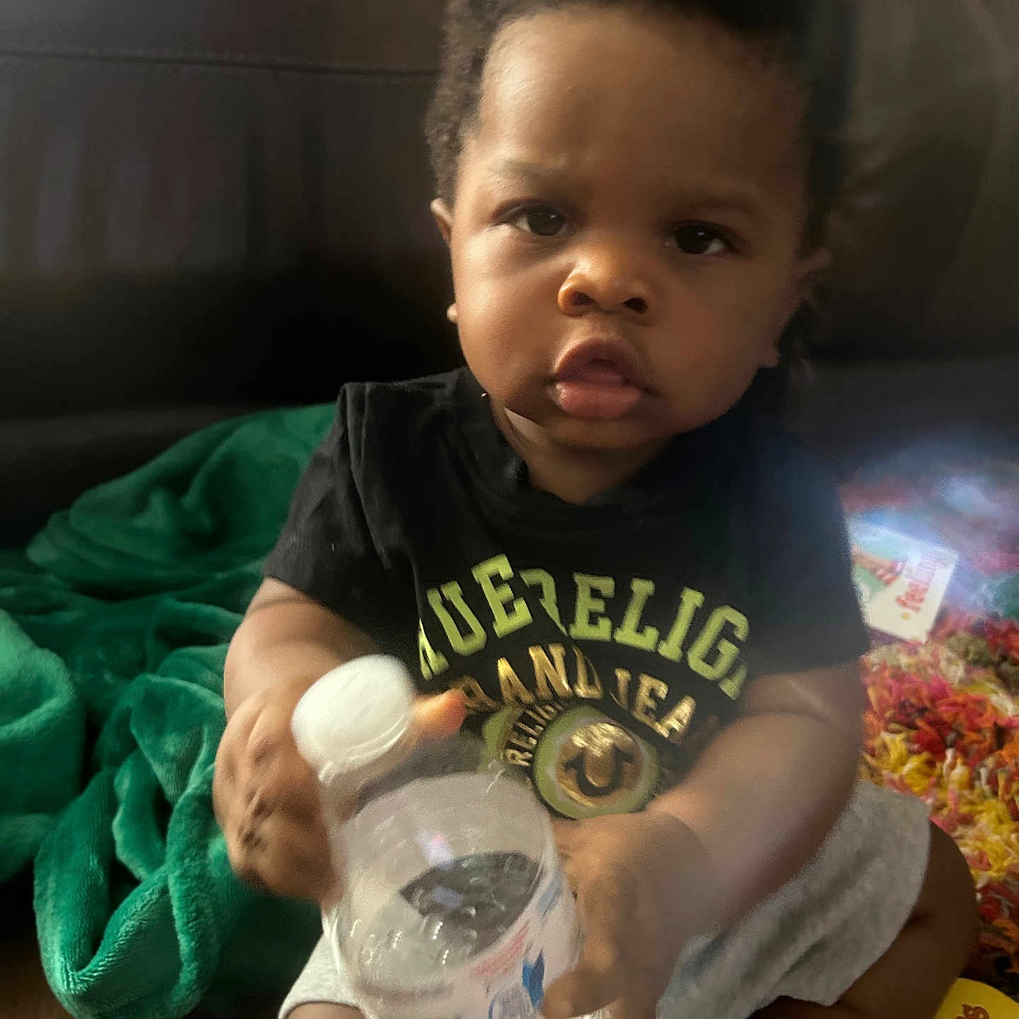 Derrick is registered to the contest to win money with this photo: black_shirt, blanket, bottle, carpet, child, clothing, colorful, curly_hair, expression, face, floor, home, indoor, person, shorts, sitting, socks, toddler, toy, water_bottle
