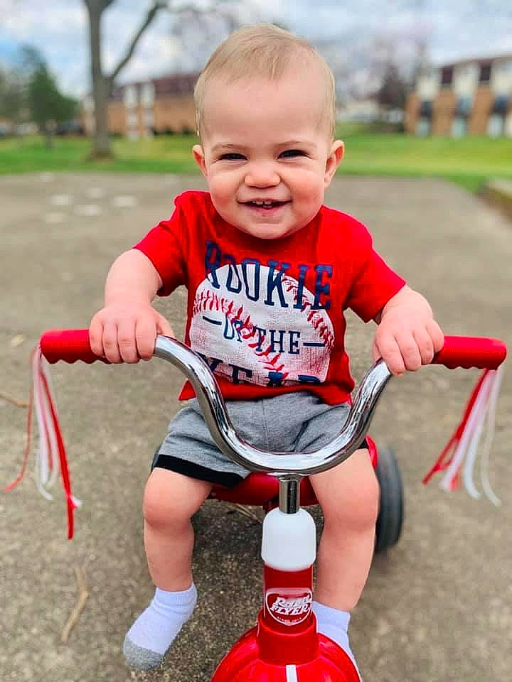 Camdyn is registered to the contest to win money with this photo: baby_playing_with_toys, baby_toddler_clothing, child, face, fun, grass, happy, human_body, human_leg, joy, knee, person, plant, recreation, red, riding_toy, shorts, smile, tire, toddler