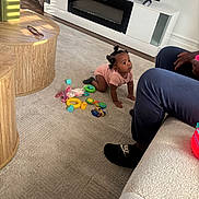 Zehlani is registered to the contest to win money with this photo: toddler, child, crawling, toys, carpet, living_room, furniture, television, person, footwear, nike, slippers, wooden_table, pink_clothing, natural_light, shadow, adult, couch, home_interior, curious