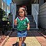 child, girl, toddler, outdoor, sunny, portrait, soccer_jersey, green_shirt, denim_skirt, red_shoes, curly_hair, hair_bows, standing, walkway, front_porch, house, stairs, stroller, garden, shadow