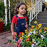 child, toddler, girl, portrait, outdoors, garden, flowers, colorful_flowers, plants, porch, stairs, house, overalls, red_shirt, hair_bow, standing, looking_down, curious, daylight, residential_exterior