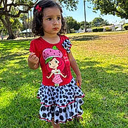 Nayleen is registered to the contest to win money with this photo: child, toddler, girl, portrait, outdoor, park, grass, sunny, dress, red_shirt, cartoon, polka_dots, skirt, barefoot, flower_hair_clip, dark_hair, standing, serious_expression, trees, palm_tree