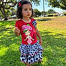 child, toddler, girl, portrait, outdoor, park, grass, sunny, dress, red_shirt, cartoon, polka_dots, skirt, barefoot, flower_hair_clip, dark_hair, standing, serious_expression, trees, palm_tree