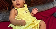 Jazlynn is registered to the contest to win money with this photo: baby, child, infant, portrait, sitting, lying_down, yellow_dress, pink_bow, sandals, smiling, curious, indoors, bed, pillows, blanket, red_bedding, brown_pillow, dark_skin, short_hair, casual_photo