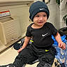 beanie, black_clothing, blanket, casual_wear, changing_table, child, cute, footwear, hat, indoor, nike, person, plant, portrait, public_restroom, smiling, sneakers, tile_wall, toddler, young_child