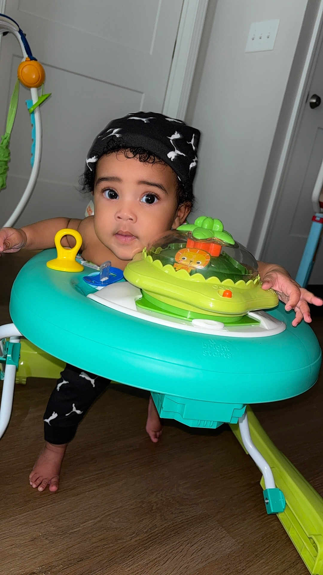 Blaze is registered to the contest to win money with this photo: baby, infant, toddler, child, walker, activity_center, toy, hat, big_eyes, curly_hair, surprised_expression, barefoot, wooden_floor, door, door_handle, indoor, playtime, bright_colors, hand, face