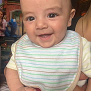 Leo is registered to the contest to win money with this photo: baby, smile, bib, striped_clothing, indoor, television, toy, hand, person, face, happy, watch, child, infant, furniture, bed, room, skin, holding, casual