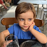 Hugo a rejoint le concours — aidez-le/la à gagner de superbes lots ! child, blue_shirt, glass, water, table, plate, chair, face, person, indoor, curious, young, short_hair, skin, hand, seated, restaurant, reflective, expression, daylight