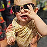 baby, child, sunglasses, hand, person, holding, smile, playful, face, clothing, patterned, adult_hands, indoor, store, blurred_background, cute, fun, head, skin, infant