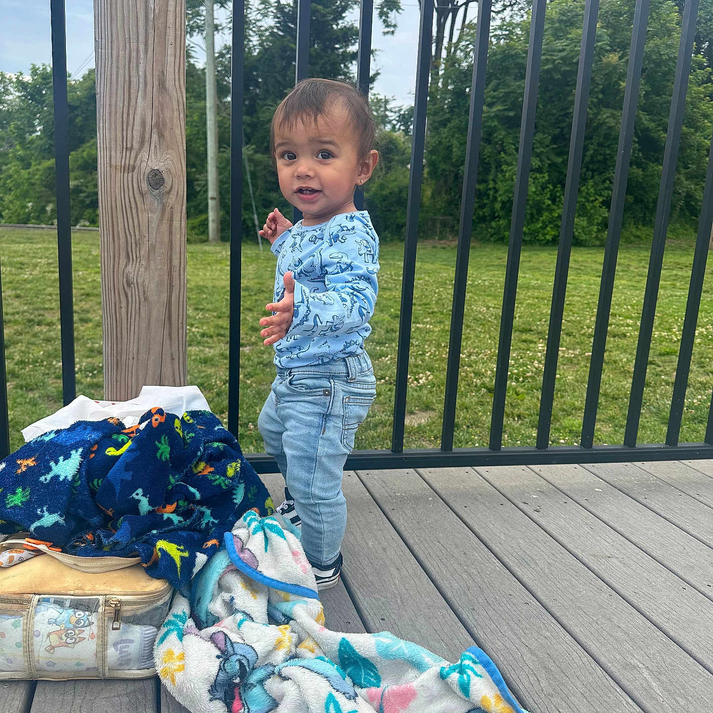 Koa joined the competition — help win amazing prizes! bag, blanket, child, clothing, daylight, deck, expression, footwear, grass, greenery, jeans, nature, outdoor, person, railing, shirt, standing, toddler, trees, wood