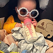 Charlie is registered to the contest to win money with this photo: baby, bottle, milk, sunglasses, car_seat, blanket, toy, wooden_sign, 4_months, clothing, infant, cute, child, feeding, accessory, seatbelt, soft_toys, portrait, indoor, headwear