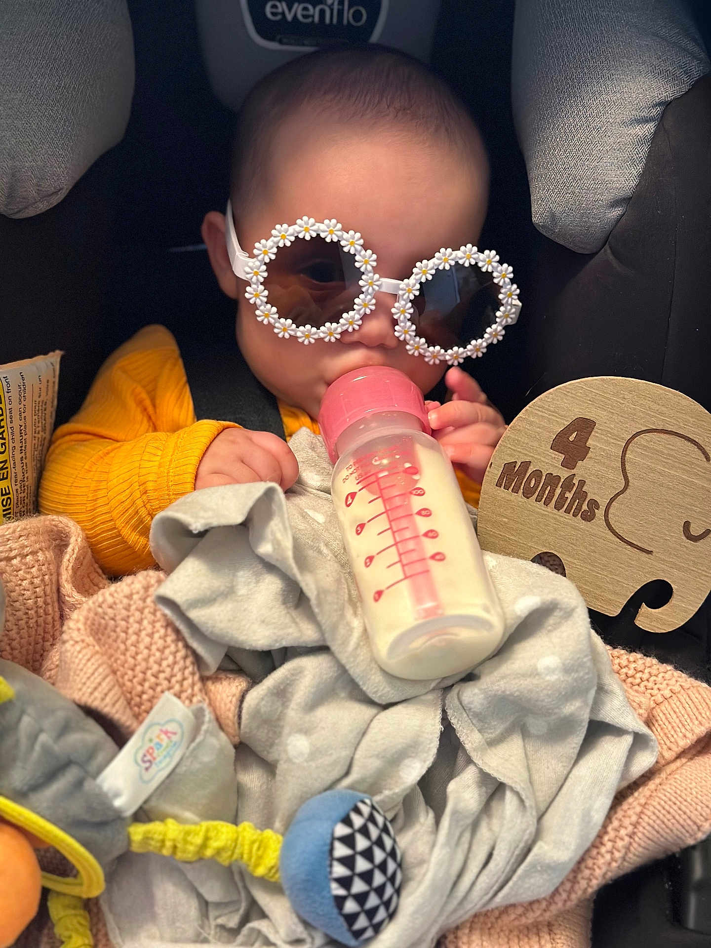 Charlie is registered to the contest to win money with this photo: baby, bottle, milk, sunglasses, car_seat, blanket, toy, wooden_sign, 4_months, clothing, infant, cute, child, feeding, accessory, seatbelt, soft_toys, portrait, indoor, headwear