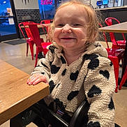 Lilyahna is registered to the contest to win money with this photo: background, black_and_white, casual_clothing, child, cozy, face, fleece_jacket, hair, happy, highchair, indoor, neon_sign, red_chairs, restaurant, seated, smiling, table, toddler, wall_art, wooden_table