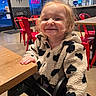 background, black_and_white, casual_clothing, child, cozy, face, fleece_jacket, hair, happy, highchair, indoor, neon_sign, red_chairs, restaurant, seated, smiling, table, toddler, wall_art, wooden_table