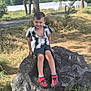 child, boy, rock, outdoor, grass, tree, smile, shorts, shirt, sneakers, nature, sunlight, casual, playful, park, daylight, young, happy, portrait, leisure