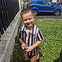 child, boy, smiling, grass, flower, fence, car, shorts, shirt, outdoor, daylight, sidewalk, parked_car, happy, nature, building, sneakers, greenery, summer, portrait