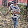 child, boy, sunglasses, smiling, outdoor, grass, fence, shirt, jeans, shoes, happy, casual_clothing, nature, portrait, walking, daylight, young, person, fashion, summer