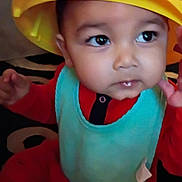 Ezra is registered to the contest to win money with this photo: baby, child, hat, tonka, yellow_hat, bib, red_clothing, indoor, carpet, face, eyes, cute, infant, person, young_child, sitting, curious, closeup, headwear, portrait