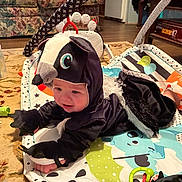 Levi joined the competition — help win amazing prizes! baby, child, costume, skunk_costume, play_mat, toy, floor, carpet, bottle, indoor, living_room, furniture, person, cute, infant, black_clothing, smiling, face, hand, feet