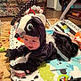 baby, child, costume, skunk_costume, play_mat, toy, floor, carpet, bottle, indoor, living_room, furniture, person, cute, infant, black_clothing, smiling, face, hand, feet