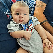 Levi is registered to the contest to win money with this photo: baby, infant, person, child, blue_eyes, onesie, american_flag, arm, tattoo, casual_clothing, indoor, seated, curious_expression, chubby, holding, adult, background, blurred, skin, closeup