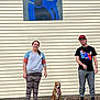 Lizzy joined the competition — help win amazing prizes! people, dog, leash, sidewalk, building, sign, wall, casual_clothing, woman, man, sneakers, hat, pants, shirt, outdoor, pet, tongue_out, smile, daytime, companionship