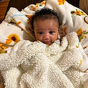 Genesis is registered to the contest to win money with this photo: baby, infant, child, blanket, floral_pattern, sunflower, curly_hair, big_eyes, smile, portrait, indoors, cozy, soft_texture, furry_blanket, wrapped, cute, eye_contact, sofa, wood_floor, cardboard_box