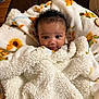 baby, infant, child, blanket, floral_pattern, sunflower, curly_hair, big_eyes, smile, portrait, indoors, cozy, soft_texture, furry_blanket, wrapped, cute, eye_contact, sofa, wood_floor, cardboard_box