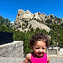 child, toddler, pink_dress, curly_hair, smiling, mount_rushmore, rock_sculpture, blue_sky, outdoor, tourist, landmark, granite, nature, trees, sunlight, portrait, happy, daytime, travel, vacation