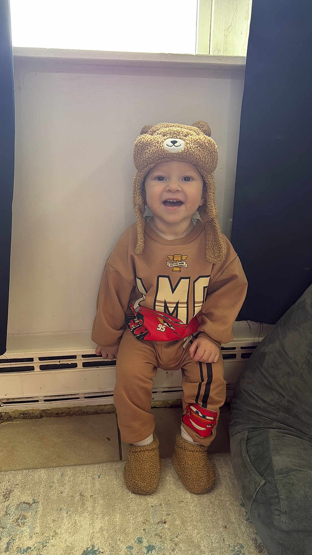 Henry joined the competition — help win amazing prizes! toddler, child, hat, slippers, brown_clothing, indoor, window, smiling, sitting, car_character, cozy, floor, carpet, radiator, curtains, cute, face, happy, person, toy_theme