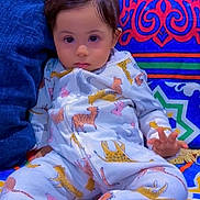 Moh is registered to the contest to win money with this photo: baby, child, pajamas, animal_print, patterned_clothing, colorful_background, couch, soft_furniture, indoor, cute, portrait, sitting, young_child, face, hand, expression, comfort, relaxed, home, cozy