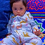baby, child, pajamas, animal_print, patterned_clothing, colorful_background, couch, soft_furniture, indoor, cute, portrait, sitting, young_child, face, hand, expression, comfort, relaxed, home, cozy
