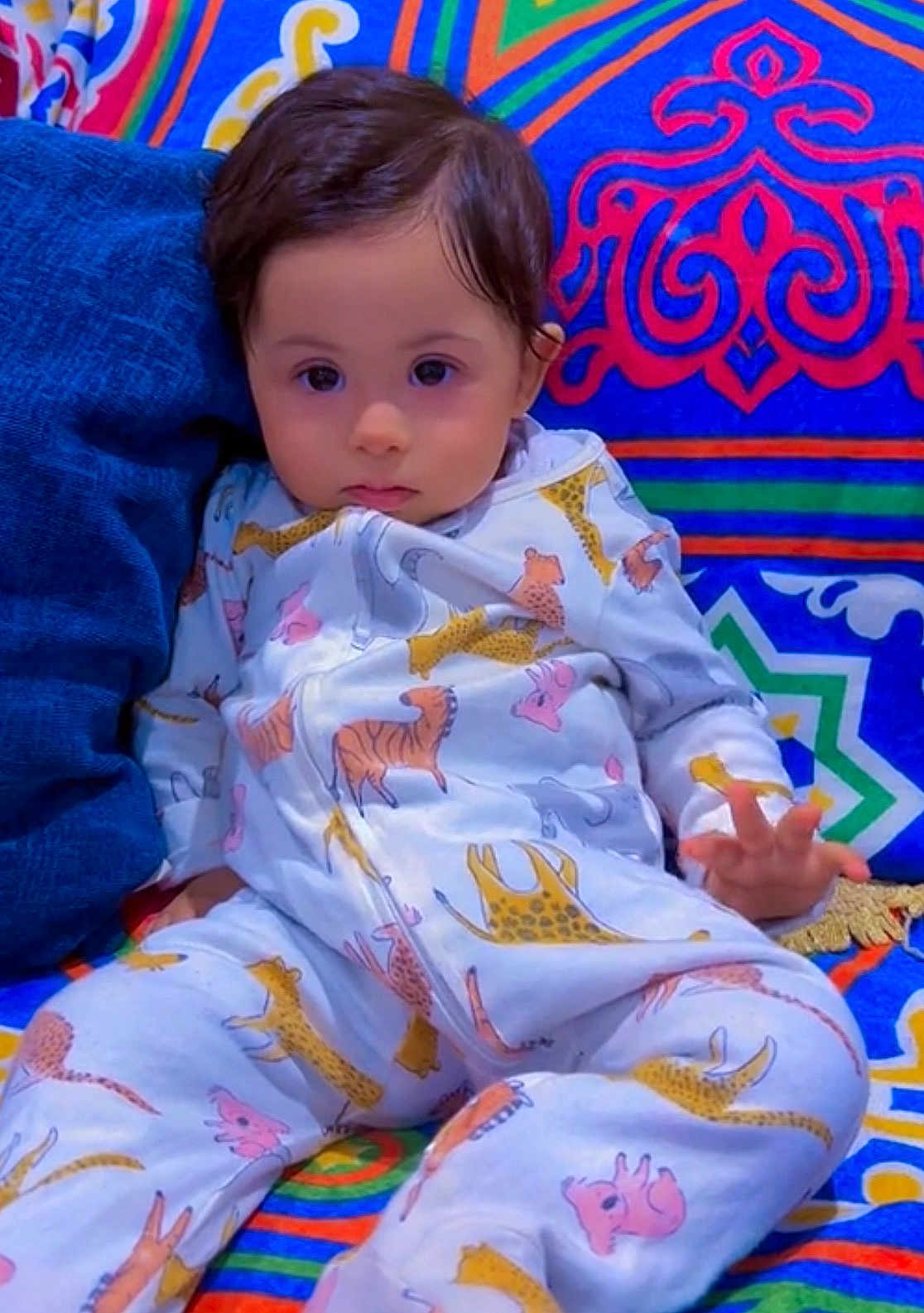 Moh is registered to the contest to win money with this photo: baby, child, pajamas, animal_print, patterned_clothing, colorful_background, couch, soft_furniture, indoor, cute, portrait, sitting, young_child, face, hand, expression, comfort, relaxed, home, cozy