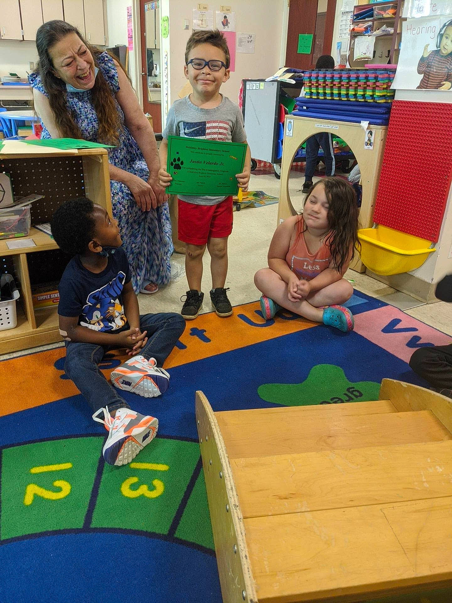 Justin is registered to the contest to win money with this photo: academic_institution, art, chair, child, class, event, flooring, fun, joy, kindergarten, learning, leisure, person, play, recreation, room, sharing, shorts, smile, t_shirt