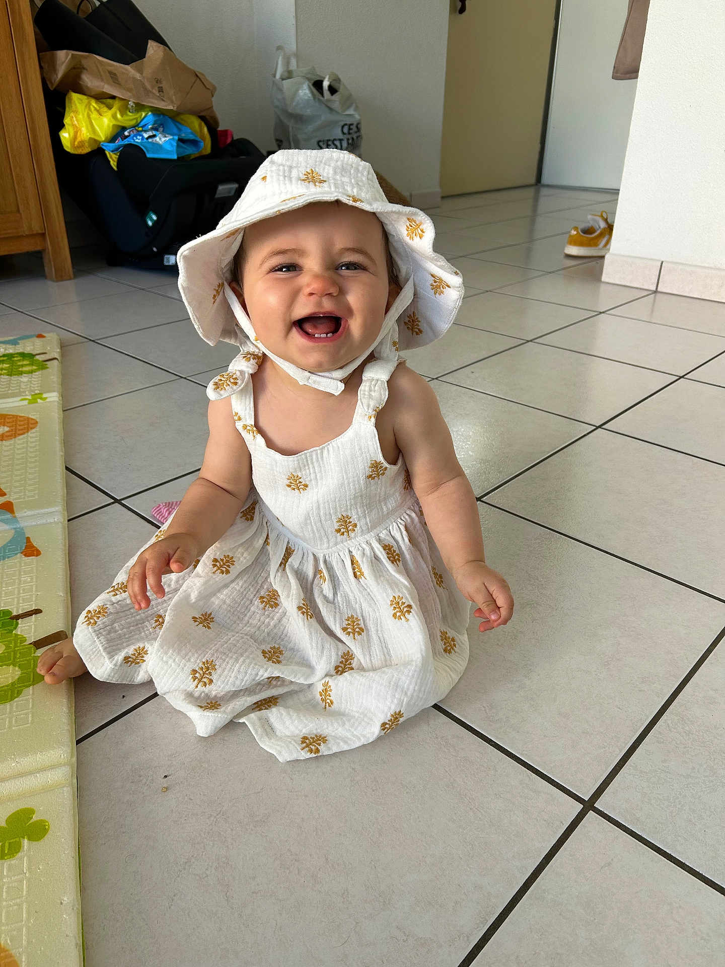 Naëlya a rejoint le concours — aidez-le/la à gagner de superbes lots ! baby, child, smiling, happy, dress, hat, floor, tile, indoor, person, cute, sitting, childhood, playmat, home, young, infant, apparel, headwear, joy
