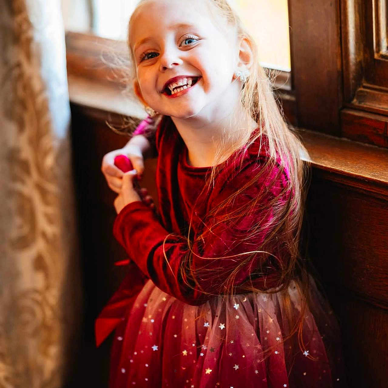 Ivie-Alice is registered to the contest to win money with this photo: child, curtain, cute, earrings, fashion, girl, happy, indoor, light, long_hair, portrait, posing, red_dress, smile, sparkle, stars, sunlight, velvet, window, wooden