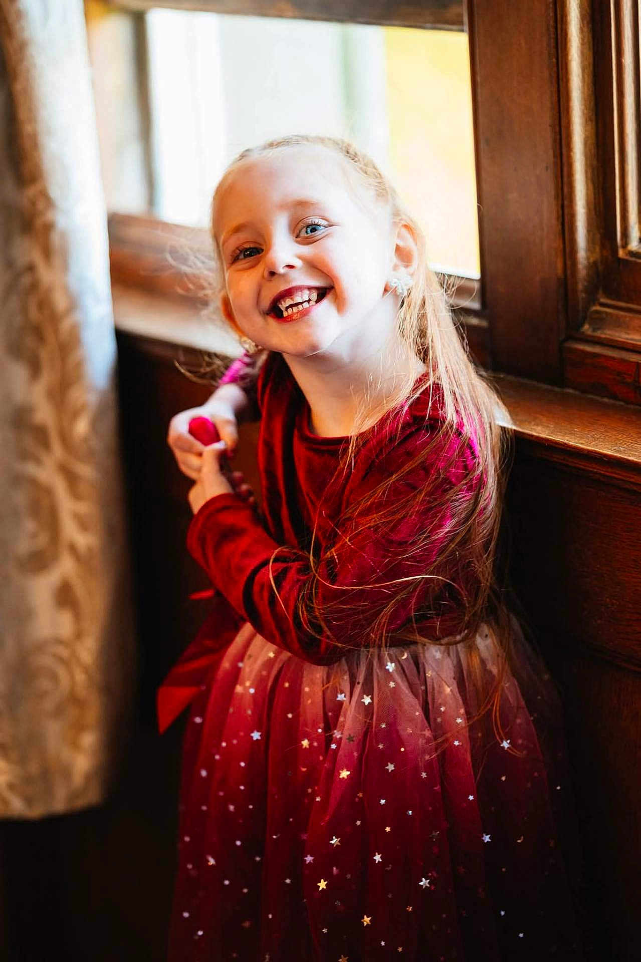 Ivie-Alice is registered to the contest to win money with this photo: child, girl, smile, red_dress, velvet, stars, long_hair, window, wooden, curtain, indoor, portrait, happy, light, sunlight, sparkle, earrings, posing, cute, fashion
